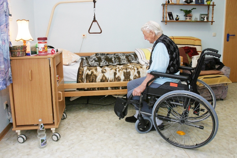 Seniore Person im Rollstuhl vor seinem Seniorenbett mit Halterung.