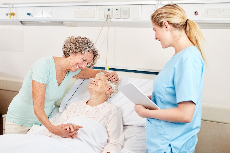 Eine Pflegekraft in einem hellblauen Kittel steht in einem Patientenzimmer einer Seniorin. Die Seniorin liegt in ihrem Bett, neben ihr steht eine Angehörige, die ihr liebevoll über den Kopf streichelt.