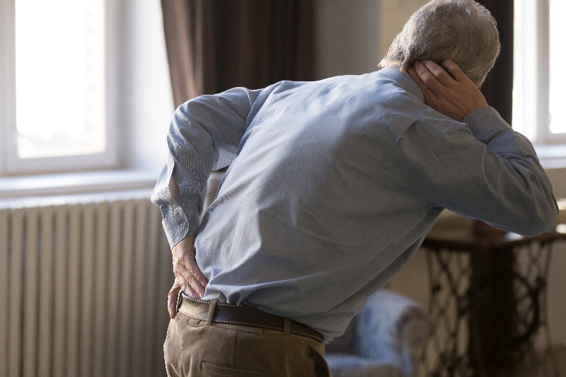 Mann mit grauen Haaren hält sich den Rücken und lehnt sich mit dem anderen Ellenbogen an. Symbolbild für Erkrankungen des Bewegungsapparats