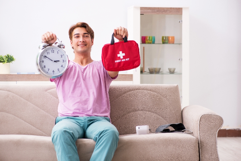 Junger Mann sitzt auf einem hellen Sofa und hält einen retro weißen Wecker in der einen und einen roten Notfallkoffer in der anderen Hand. Der Mann trägt ein rosa T-Shirt und eine salbeifarbene Hose. Neben ihm auf dem Sofa liegt ein Blutmessgerät. Hinter ihm zu sehen ist ein Regal mit bunten Vasen und eine grüne Zimmerpflanze.