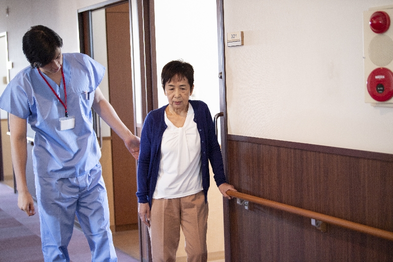 Dame mit Gangschwierigkeiten hält sich an einer Stange und wird von Pfleger begleitet.