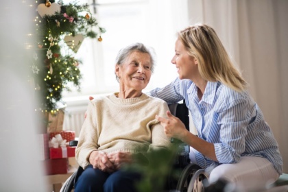 Eine Angehörige in einer blauen Bluse kümmert sich zu Weihnachten um eine Seniorin im Rollstuhl. Im Hintergrund steht ein Weihnachtsbaum mit Festtagsdekoration.