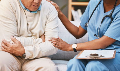 Eine junge Ärztin in blauem Kittel hat ihre Hände beruhigend auf den Arm und die Schulter eines Senioren gelegt. Der Senior trägt einen cremefarbenen Pullover und hält sich mit beiden Armen um den Bauch fest. Die beiden sitzen auf einem hellgrauen Sofa. Im Hintergrund ist die Wand dunkelgrün gestrichen und ein Holzregal mit Büchern ist zu erkennen.