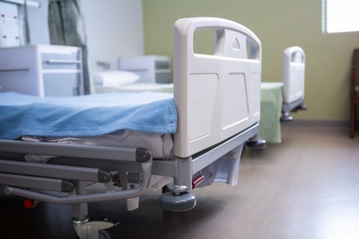 Im Fokus stehen zwei Krankenhausbetten, eins im Vorder- und eins im Hintergrund. Die Wand ist Zitronengelb gestrichen. Auf dem Bett im Vordergrund liegt eine blaue Unterlage, auf dem Bett im Hintergrund eine grüne. Neben den Betten steht jeweils ein weißer Nachttisch mit grauen Schubladen. Zwischen den Betten kann ein dunkelgrüner Vorhang gespannt werden.