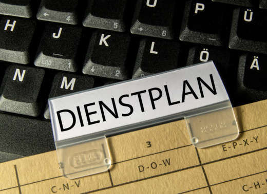 Auf einer schwarzen Tastatur mit weißen Ziffern liegt ein hellbraunes Dokument, an dem eine Klammer geheftet ist. Auf der Klammer steht in schwarzer Schrift auf weißem Papier das Wort „Dienstplan“.