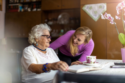 Eine Seniorin sitzt an einem Tisch. Auf dem Tisch steht eine Tasse mit Blumenmuster, eine Orchidee in einem rosa Blumentopf und zwei Fernbedienungen. Auf dem Tisch ist ein weißes Spitzentischtuch ausgebreitet. Die Senioren trägt ein weißes Oberteil, eine blaue Kette und ein blaues Armband. Sie trägt hellblaue Ohrringe und eine schwarze Brille. Auf dem Tisch in ihrer Hand ist ein aufgeschlagenes Buch gelegt. Eine junge Frau in lila Oberteil und schwarzer Hose beugt sich lächelnd zu der Seniorin herunter und hat ihre rechte Hand auf die linke Schulter der Seniorin gelegt. Im Hintergrund ist eine hölzerne Wohnwand mit mehrere Dekoartikeln.