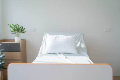 Im Zentrum des Bildes steht ein sauberes Krankenhausbett mit Kissen und Laken. Links daneben steht eine Kommode, auf der eine Topfpflanze steht. Die oberste Schublade ist schwarz, der Rest Holzfarben. Vor der Kommode steht eine weitere Topfpflanze. Die Wand ist weiß gestrichen, der Boden ist aus Holz.