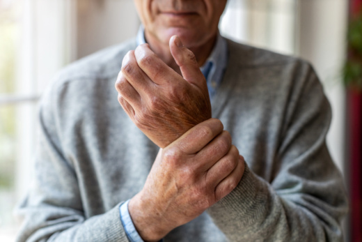 Ein Senior in grauem Pullover hält mit seiner rechten Hand das Handgelenk seiner linken Hand in die Kamera. Sein Mund ist ernst. Der Hintergrund zeigt unscharf ein Fenster.