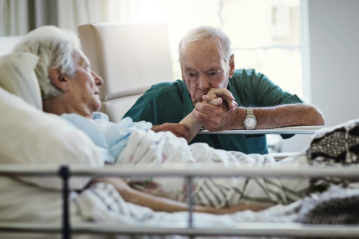 Ein Senior in dunkelgrünem T-Shirt sitzt am Bett einer Seniorin. An seinem Handgelenk trägt er eine goldene Armbanduhr. Er hält die Hand der Seniorin und küsst ihre Hand. Die Seniorin schaut ihn an. Sie trägt ein hellblaues T-Shirt. An ihrem Bett sind Bettgitter befestigt. Beide haben einen ernsten Gesichtsausdruck. Im Hintergrund ist ein Fenster und ein beiger Sessel zu erkennen. Die Bettdecke der Seniorin ist mit einem Blumenmuster bedruckt.