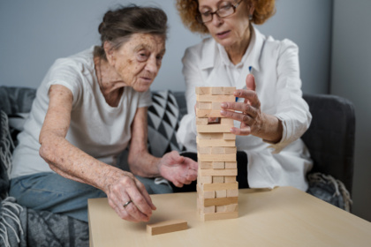 Auf einem Beistelltisch steht ein Jenga-Turm. Eine Seniorin und eine Ärztin sitzen auf einem Sofa davor und spielen Jenga. Beide haben einen ernsten Gesichtsausdruck. Die Seniorin trägt ein weißes T-Shirt und eine blaue Jeans. An ihrer Hand trägt sie einen Ring. Die Ärztin trägt einen weißen Kittel und hat lackierte Fingernägel.