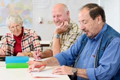 Drei Senioren sitzen in einem Klassenzimmer an einem Tisch. Sie halten Stifte in der Hand und haben aufgeschlagene Notizbücher vor sich liegen. Der Senior im Zentrum trägt ein blau-weiß kariertes Hemd und eine Jeansweste. Er schaut in sein Notizbuch. Der Senior neben ihm trägt ein grün-kariertes Hemd und schaut in das Notizbuch des Seniors neben ihm. Ganz links sitzt ebenfalls eine Seniorin in einem rot-weiß kariertem Hemd. Sie trägt eine Brille und schaut zu den beiden Senioren neben ihr.