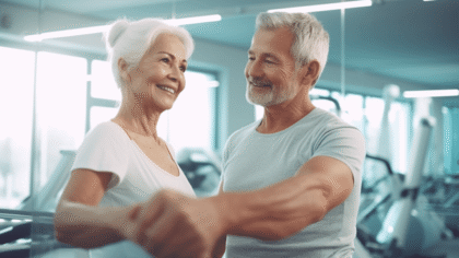 Zwei ältere Menschen stehen gemeinsam in einem Fitnessstudio. Der Mann auch der rechten Seite unterstützt die Frau auf der linken Seite bei einer körperlichen Aktivität. Beide lächeln zufrieden. Die Frau trägt ein weißes T-Shirt, der Mann trägt ein graues T-Shirt. Im Hintergrund sind weitere Trainingsgeräte zu erkennen.