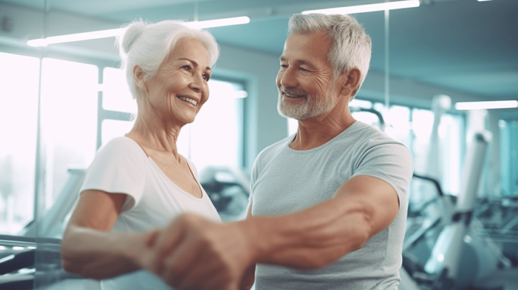 Zwei ältere Menschen stehen gemeinsam in einem Fitnessstudio. Der Mann auch der rechten Seite unterstützt die Frau auf der linken Seite bei einer körperlichen Aktivität. Beide lächeln zufrieden. Die Frau trägt ein weißes T-Shirt, der Mann trägt ein graues T-Shirt. Im Hintergrund sind weitere Trainingsgeräte zu erkennen.