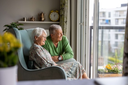Zwei Rentner befinden sich in einem Wohnzimmer einer Seniorenresidenz. Die Seniorin sitzt in einem Sessel, der Senior kniet neben ihr. Beide lachen und schauen aus der Balkontür nach draußen.