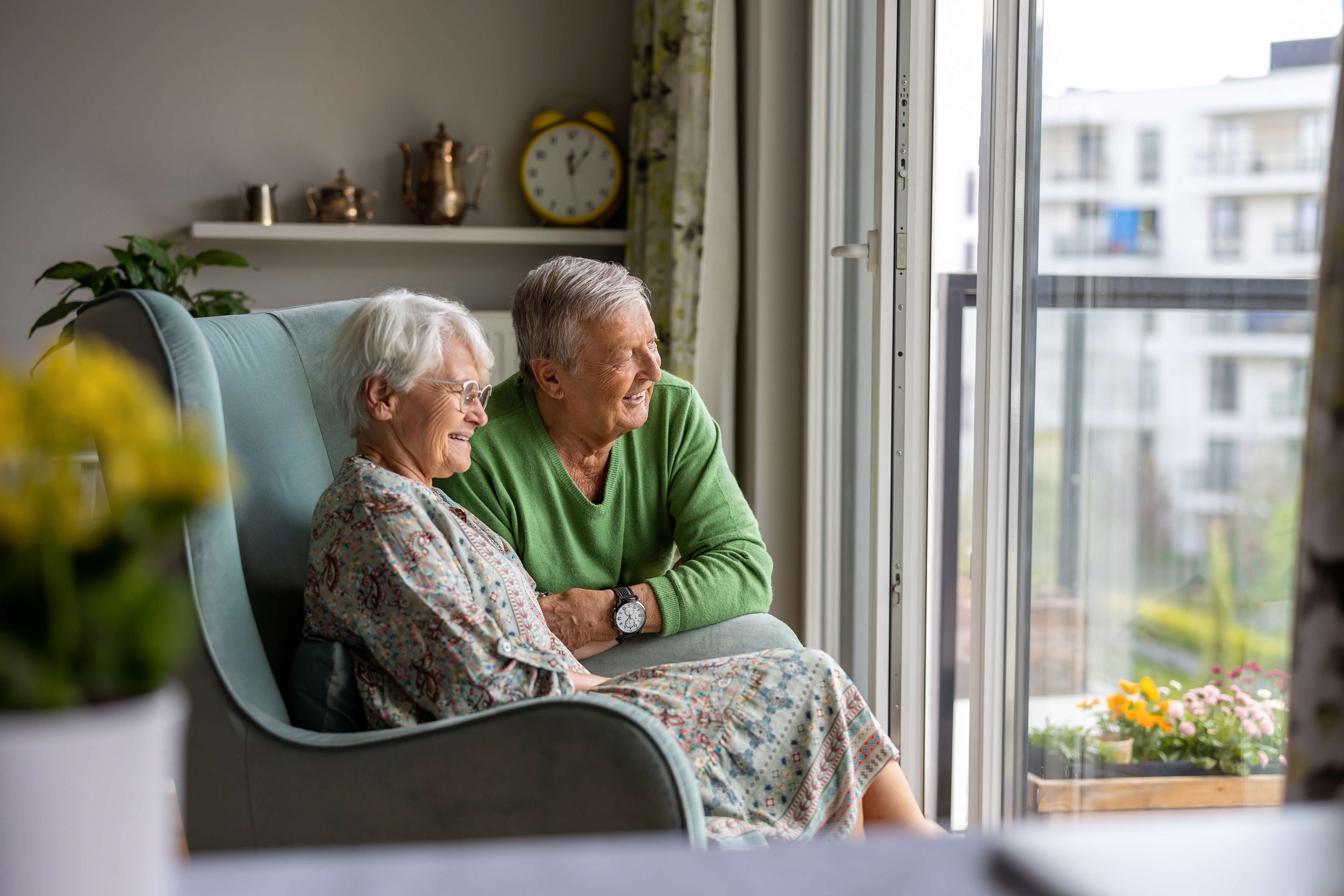 Zwei Rentner befinden sich in einem Wohnzimmer einer Seniorenresidenz. Die Seniorin sitzt in einem Sessel, der Senior kniet neben ihr. Beide lachen und schauen aus der Balkontür nach draußen.