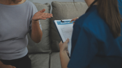 Eine Seniorin gestikuliert mit einer Pflegerin, welche eine Anamnese auf einem Klemmbrett zur Inkontinenz durchführt.