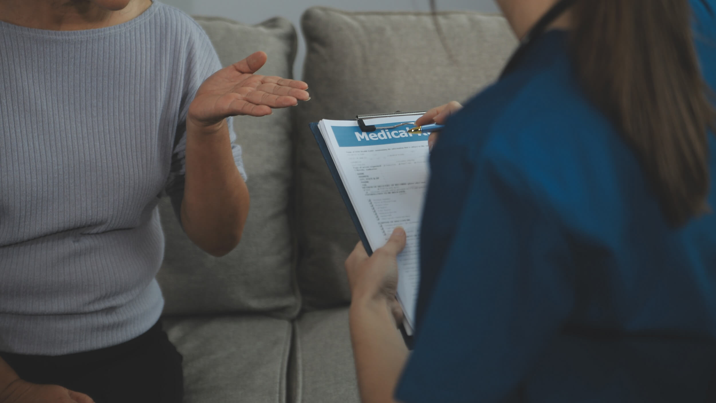 Eine Seniorin gestikuliert mit einer Pflegerin, welche eine Anamnese auf einem Klemmbrett zur Inkontinenz durchführt.