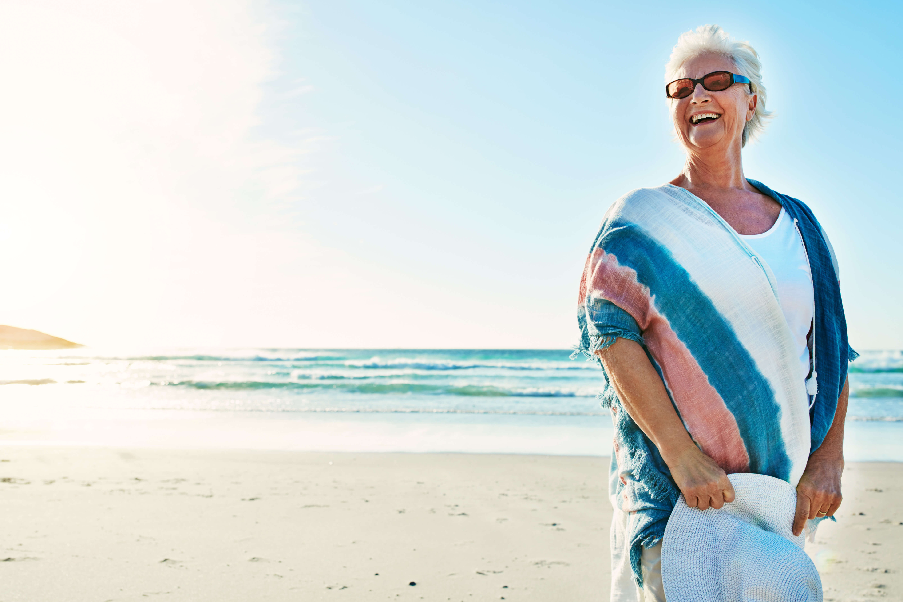 Eine Seniorin in luftigen Sommeroutfit trägt Sonnenbrille und Hut und lacht fröhlich am Strand.