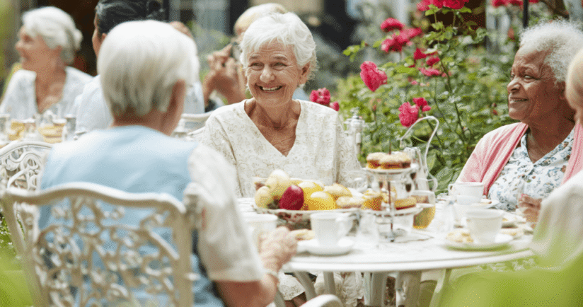 Gruppe von vier Seniorinnen sitzt lächelnd am Gartentisch.