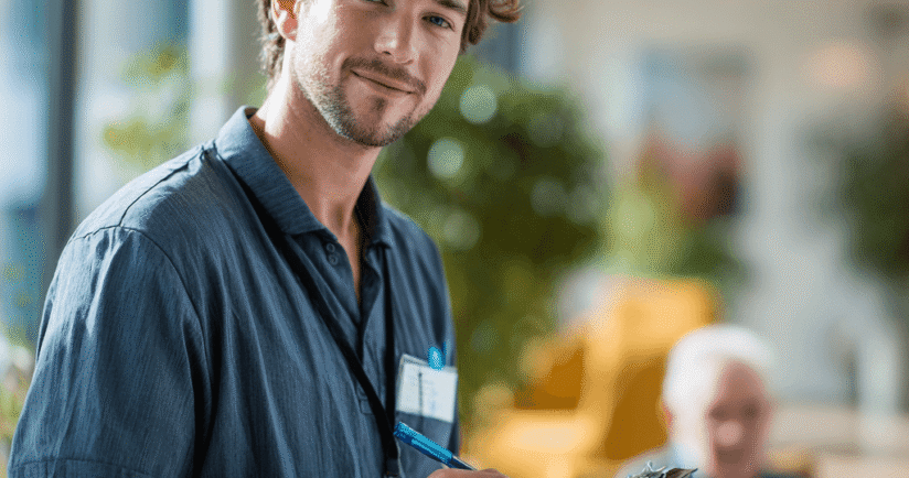 Junger Pfleger in blauem Kasack schreibt etwas auf ein Klemmbrett. Im Hintergrund ist ein alter Pflegepatient zu sehen.