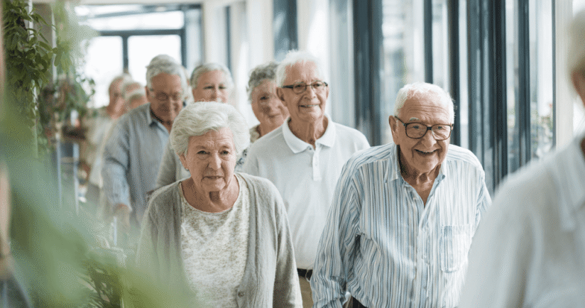 Gruppe an Senior*innen geht zu Fuß, am Krückstock oder mit einem Rollator über einen hellen Gang einer Altenpflegeeinrichtung.