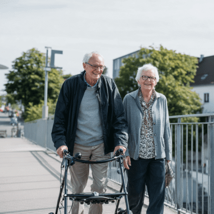 Ein gepflegtes Seniorenpaar ist bei gutem Wetter draußen. Die lächeln sich an. Der Herr hat einen Rollator.
