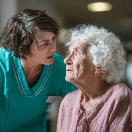 Pflegefachkraft in Arbeitskleidung beugt sich in einer Pflegeeinrichtung über eine hochbetagte Frau; die Pflegekraft wirkt angespannt und laut, die Seniorin traurig und eingeschüchtert, Hintergrund hell und unscharf.