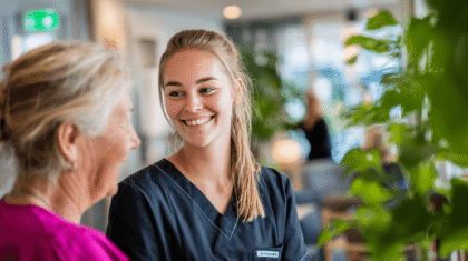 Junge blonde Pflegerin in der Ausbildung, lächelt eine andere Person an.