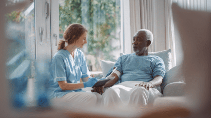 Pflegefachkraft sitzt am Bett mit Senior, der eine Wunde am Arm hat. Sie dokumentiert diese