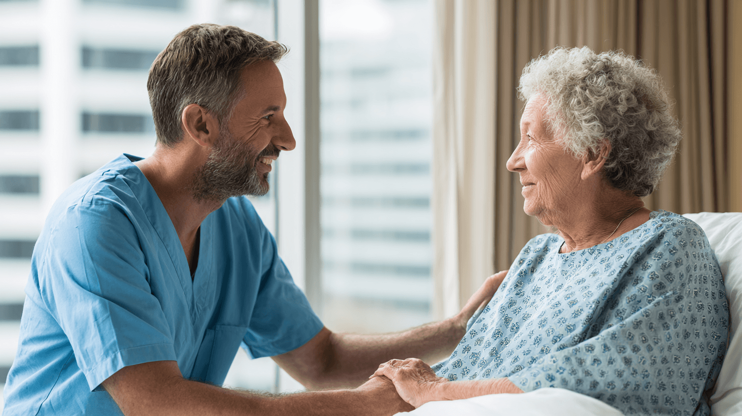Altenpfleger mit Bart und gräulichen Haaren sitzt am Bett einer senioren Pföegepatientin und berührt sie vorsichtig, Symbolbild für basale Stimulation