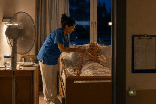 Pflegekraft bietet Seniorin im Bett ein Glas Wasser an, während der Ventilator läuft. Symbolbild für Hitzeschutz in der Nacht